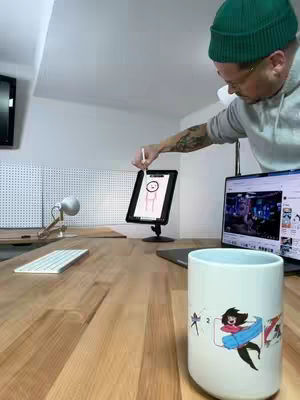 A man in a green beanie and glasses is holding a stylus and drawing on a tablet that is placed on a stand. A laptop and a white mug with illustrations are visible on a wooden desk, a keyboard and desk lamp are in the background.