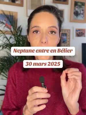 A woman with brown hair and eyes is centered in the frame with a red sweater. A white box overlay with text appears at the center of frame and bottom, above the woman's chest, with the words “Neptune entre en Bélier”. Below this, the text box continues with “30 mars 2025”. The background is a domestic interior with framed artwork hanging on the wall, partially obscured, with some of a plant visible on the left side of the woman. The woman is holding a microphone.