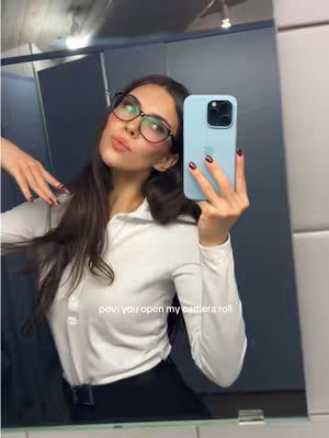 A selfie taken in a bathroom. The main subject is a person holding a light blue iPhone, their face is partially covered with the phone. The person has long brown hair, glasses, and is wearing a white shirt. The background includes a mirror, a partially visible blue wall, and a toilet structure. The lighting is moderate, with soft tones dominating the image.