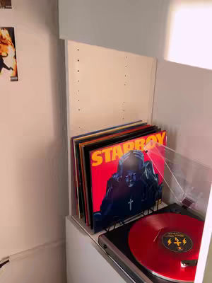 A photograph depicts a corner of a room, focusing on a stack of vinyl records. The records are in sleeves, with the prominent one featuring a bold red background and the word 'STARBOY' in large orange letters. The cover art also displays a figure wearing a mask or helmet, a cross pendant, and a small, gold cross that is illuminated. To the right of the records is a red record playing on vinyl and a black turntable. A white shelf is visible, offering space for the records and a portion of the wall, along with an adjacent wall. The lighting enhances the details.