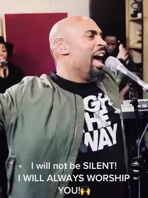 The image shows a man singing into a microphone. He's bald and wearing a black t-shirt with the words 'Give Me The Way' written on it in white. He's also wearing a green bomber jacket. The background has a red wall on the left.