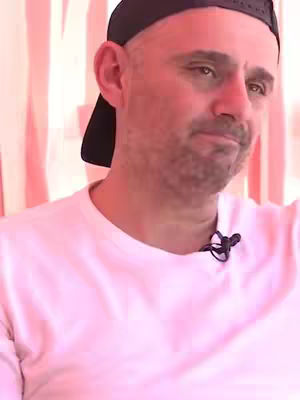 The image shows a man with a shaved head wearing a black cap and a white t-shirt. The background has vertical stripes of pink and white, indicating a possibly outdoor setting. The man is looking to his right with a neutral expression, seemingly in a candid moment.