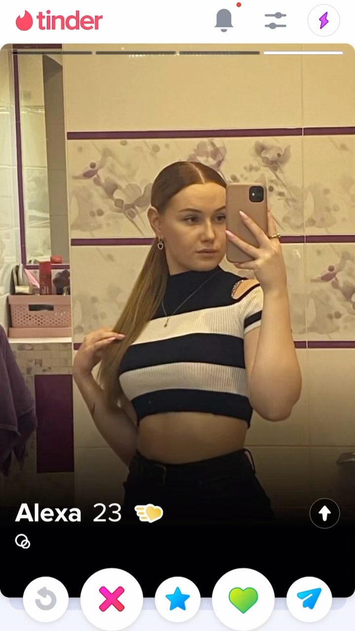 A woman takes a mirror selfie in a bathroom, holding a phone. She is wearing a black and white striped top and black pants. The background shows a tiled wall with a floral pattern. On the bottom, the interface of the app Tinder is visible indicating this is a profile picture.