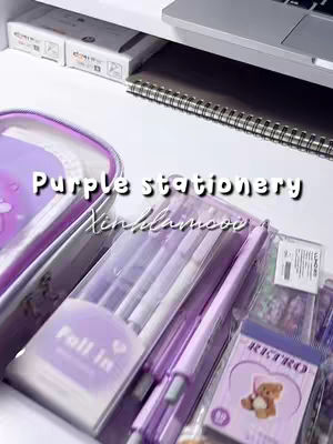 The image is a still life composition of purple-themed stationery. The foreground is dominated by a clear plastic case containing several pens, all in shades of purple. To the left is a purple pouch or pencil case. Behind it, a notebook and a portion of a laptop are visible, both on a white surface. The background includes more stationery items, such as stickers with a teddy bear illustration, and more pens. The lighting is bright and appears to be from above, highlighting the items' colors and textures.