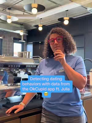 A woman with curly brown hair and red-rimmed glasses stands in a kitchen or bar setting. She has her hand raised to her chin as if deep in thought. A light blue banner hovers mid-frame; it has white text and a smiling emoji icon.