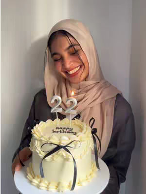 A young woman in a hijab is smiling, holding a birthday cake with the number "22" candles. The cake has white frosting and is decorated with black ribbons. The background is a light-colored wall.