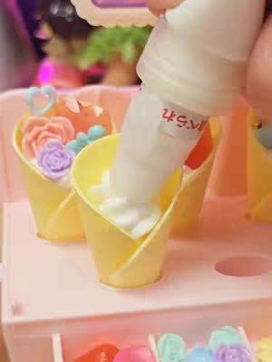 A close-up shot of a pink toy crepe wagon.  A hand holding a white nozzle dispenses white cream into a yellow crepe cone that has small toy flowers and decorations in it.
