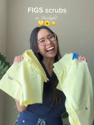 A smiling woman in glasses holds up two folded scrubs that are light yellow, likely for a product promotion. The background is a neutral color, and text overlayed on the image says FIGS scrubs in Sunlight, with emojis.