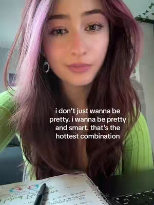 A close-up shot of a young woman with long wavy auburn hair with pink highlights. She is looking at the camera with a subtle smile. She is wearing a light green long-sleeved top and delicate hoop earrings. The text overlay reads: i don't just wanna be pretty. i wanna be pretty and smart. that's the hottest combination