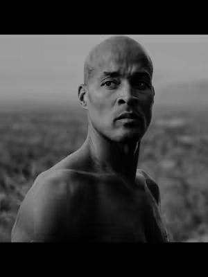 A black and white portrait-style image featuring a bald man with a stern facial expression, looking directly at the camera. The background appears blurred, suggesting a natural outdoor setting.