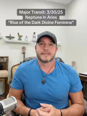 A man wearing a baseball cap and a blue t-shirt is seated in a room with a microphone below the shot. There is text at the top of the frame. The background includes a bookshelf and ornaments.