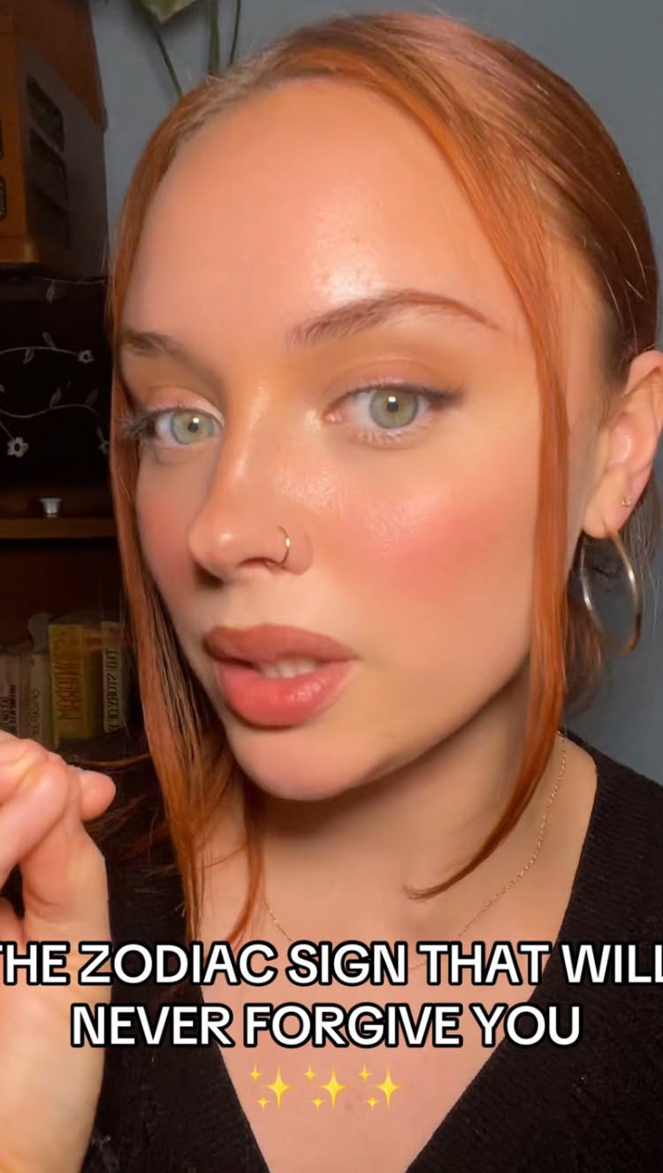 A close-up shot of a fair-skinned woman with auburn hair, centered on her face. She has a gentle expression with slightly parted lips. The video layers include text that reads, "THE ZODIAC SIGN THAT WILL NEVER FORGIVE YOU" with a background that shows a room setting that reflects the woman's personal style and is out of focus.