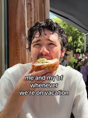 A man is shown taking a large bite from a pastry topped with a green garnish, likely pesto. The text overlay reads 'me and my bf whenever we're on vacation'. The background includes a glimpse of what seems to be a patio or outdoor setting with wooden structures.
