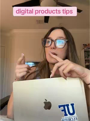 A woman with glasses is holding a laptop with a sticker on it. Above her is a light pink rectangle with the text 'digital products tips'. She is pointing with one finger while looking at the camera. The background is a ceiling fan and the top of a wall.