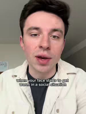A close-up shot of a young man with dark hair and a light complexion. The man is wearing a white casual jacket over a black t-shirt. The background is blurred, suggesting an indoor setting. Text overlayed on the image reads, 'when your face starts to get warm in a social situation.'