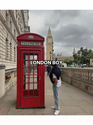 A person stands on a sidewalk in front of a red telephone booth in London. Behind them is Big Ben and other buildings under an overcast sky.
