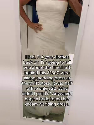 A full-length mirror shot shows a woman in a wedding dress. The dress is white, strapless, and has a fitted bodice. The woman's arms and hands are visible; one hand rests on her hip and one wears a ring. The background is a thrift store setting with racks and another garment.