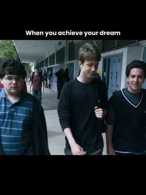 A group of three young men walking down a school hallway. The person in the middle has a semi formal black long sleeve and a black bag pack. They are walking and smiling, and the lighting is bright.