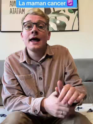 A man with glasses is speaking to the camera in front of a poster. He is wearing a brown button-down shirt and the background shows a living room setting, including a sofa. A text overlay with the words 'La maman cancer' and a Cancer zodiac symbol is visible.