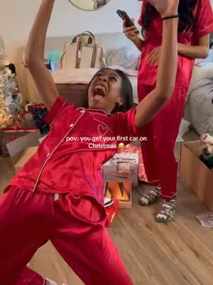 A woman, dressed in red pajamas, celebrates with arms raised and mouth open in excitement, likely receiving a gift. Behind her, another person in similar attire holds a phone, and various Christmas decorations and gift boxes are scattered in the background, suggesting a holiday surprise.