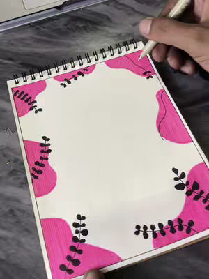 A notebook is open on a marble surface with a pen hovering over it. The notebook has an off-white page with a pink design around the edges. The pink design is made of irregular shapes with black leaf or branch-like elements spaced throughout. The notebook is open, and the hand holding the pen enters the frame.