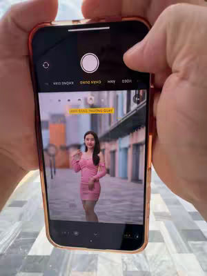 A person is holding an iPhone displaying a photo of a woman in a pink dress. The phone is the primary focus, held in hands, with the camera app open. The woman in the photo is standing on a street, with buildings in the background. The color palette is dominated by pinks, blues, and neutral tones.