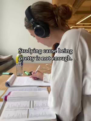 A person wearing headphones sits at a desk, studying. The person has their hair up. There are open books, papers, and a water bottle on the desk. The text overlay reads, 'Studying cause being pretty is not enough.'