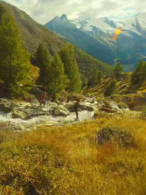 Scenic view of a mountain landscape with a river flowing through it. Two people are partially visible in the river, on the rocks, perhaps crossing. The background features a snow-capped mountain range beneath a bright sky.