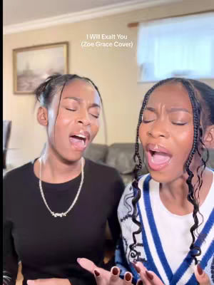 The image shows two young women, presumably twins, positioned close to each other. Both have dark hair pulled back from their faces. They are wearing makeup and appear to be in the midst of singing with their mouths open. The woman on the left is wearing a black top and a necklace, while the woman on the right is wearing a sweater with blue and white striped detailing. The background includes a neutral-colored wall, a framed picture, and a glimpse of homely interior elements.