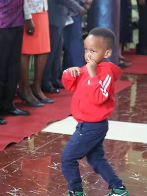 A young boy, likely a toddler, is captured mid-action, posed as if striking a martial arts stance, nose slightly scrunched. He wears a red hooded sweatshirt, blue pants, and sneakers with green accents. The background includes blurred figures, suggesting public gathering. The setting has a glossy tiled floor and portions of a red carpet.