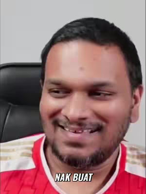 A medium shot of a man with dark hair and a slight beard, smiling and looking to the left. He's wearing a red shirt with white accents. Text overlayed at the bottom that says 'NAK BUAT'.