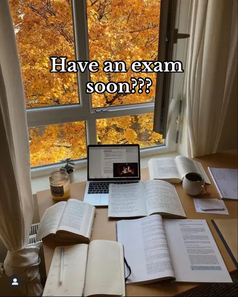 A desk by a window filled with fall foliage. The desk has several open books, a laptop displaying text, a cup, a candle, and other study materials.