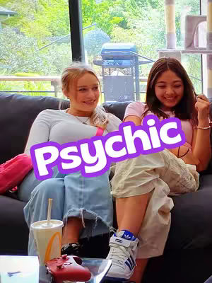 Two young women are seated close together on a dark gray couch in front of a large window. One is on the left wearing light blue jeans, a long-sleeved white shirt, and black and white sneakers. She has blonde hair and is holding a McDonald's cup. The other woman on the right has dark hair, is wearing tan shorts and a pink shirt, and is wearing white shoes with blue lines. She is smiling. A purple text banner that says 'Psychic' is over them.