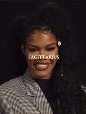 A medium shot featuring a woman with dark, voluminous curly hair and a warm smile. The woman is positioned centrally, with a dark background enhancing the focus on her. She wears a grey and black checkered blazer over a dark top. The text "SAGITTARIUS" is overlaid across her face. She has defined makeup with a focus on her eyes and glossy lips. Her smile is bright, showing white teeth, and she wears dangling earrings with an aesthetic seashell design.