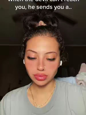 A close-up shot focuses on a person with a bun hairstyle, wearing a grey t-shirt, and small white earbuds. The person has a visible nose ring and a full-lip, with closed eyes. The background is blurred. The text overlay reads: "When the devil can't reach you, he sends you a..."