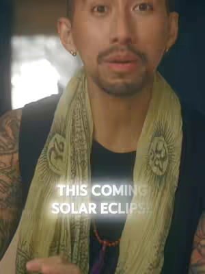 A man with a goatee and mustache looks directly at the camera. He is wearing a black tank top, a light-colored scarf, and a necklace of beads. Text overlays on the image read: “THIS COMING SOLAR ECLIPSE!”