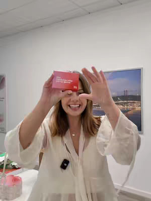 A woman is holding a red card in front of her face, framed by her hands. She is smiling. Her hair is blonde and her eyes are open. She is wearing a white blouse. The background is a plain wall and partially visible is a painting of a skyline.