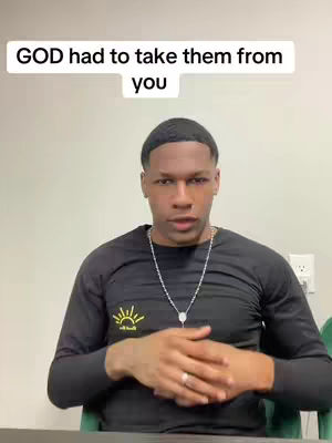 A medium shot shows a young Black man sitting in front of a light-colored wall. Text overlayed on top says, "GOD had to take them from you." The man is wearing a black long-sleeve shirt with a small yellow graphic on the left chest and what appears to be a silver necklace. He is looking directly at the camera.
