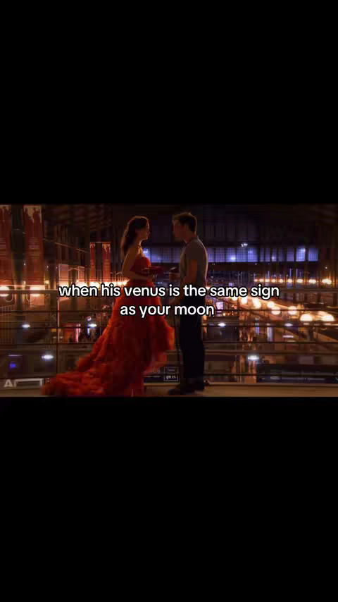 A couple is standing facing each other in a large hall. The woman on the left is wearing an elaborate red dress, while the man is wearing a gray shirt. The background is dark with some lights visible.