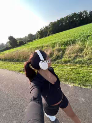 A selfie-style shot with a woman in athletic wear on a path, wearing headphones, with a green hill in the background. The sky is blue and the lighting is bright, suggesting a sunny day.