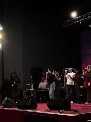 A live performance on a stage is captured. The stage is set with a red floor and several microphones and music equipment are ready. There is a group of people on the stage with microphones as well. The lighting is dim, with a main focus on the performers.