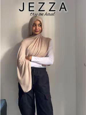 A woman wearing a hijab and fashionable clothing stands in front of a plain wall. The image has text overlayed. She has a sweet expression