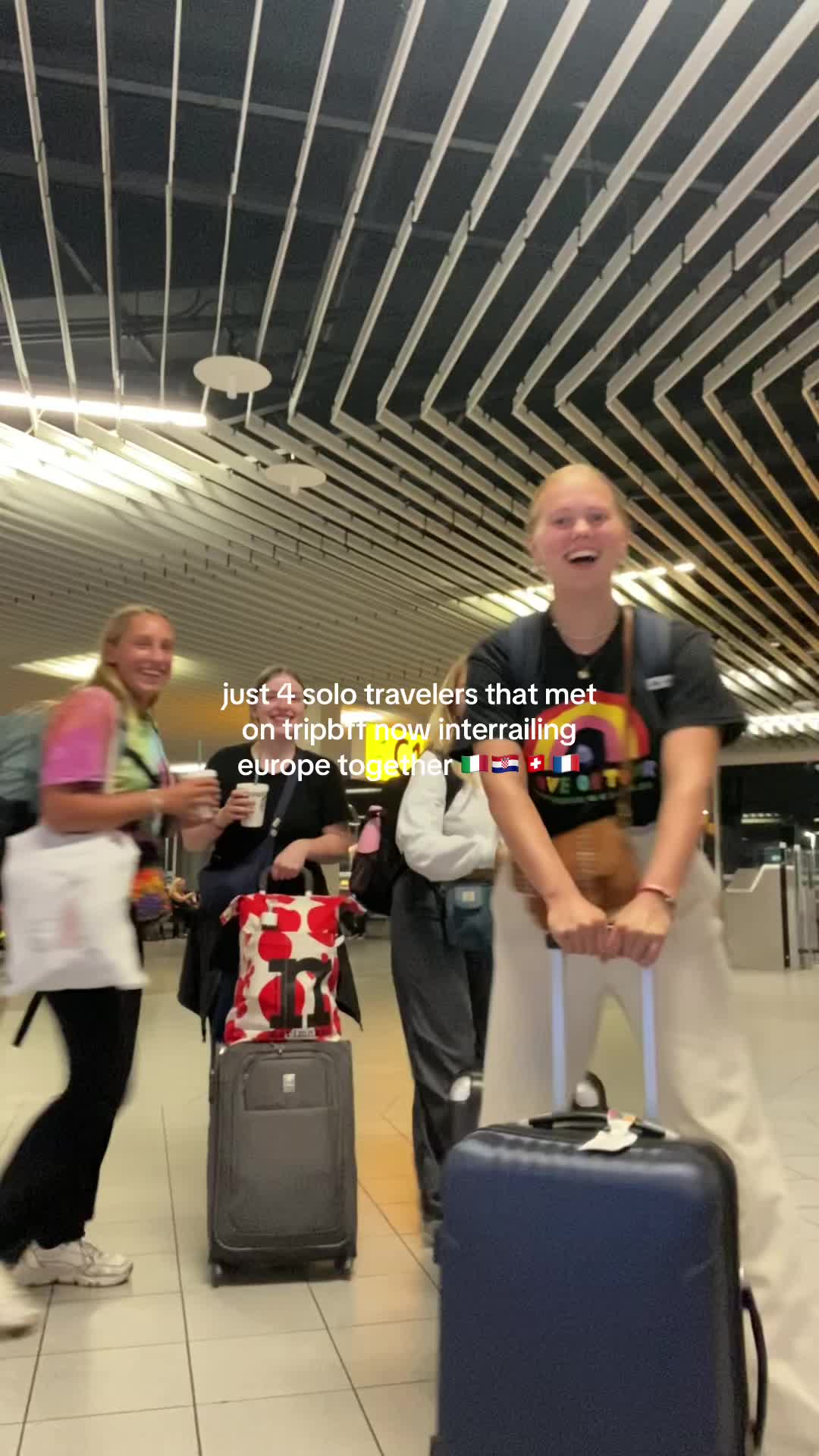 A group of four young women are at what appears to be an airport, smiling and laughing. They are pulling luggage, and there is text overlaying the image. The women are dressed in casual clothes, with colorful accents in their outfits. The background includes airport architecture with a high ceiling and long, linear lights.