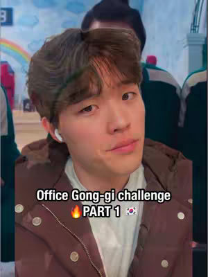 The cover image shows a young man with short, dark hair positioned in the center, slightly angled towards the left. He is wearing a brown jacket over a white shirt and has a visible earbud in his left ear. Text overlays, featuring the title "Office Gong-gi challenge PART 1" in large white text within a rectangular black box, is at the bottom. The background behind the man is somewhat blurred, and includes figures and elements associated with the Korean drama "Squid Game."