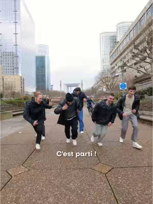 A group of young men in athletic wear run towards the camera on a paved walkway in an urban setting. Buildings and a large architectural structure are visible in the background. Text near the bottom reads 'C'est parti !'