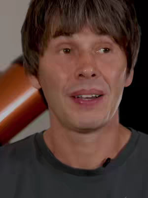 A close-up shot of a man with brown hair and a gray t-shirt. He is looking slightly to the side with a neutral expression. Behind him is an out-of-focus telescope. The lighting is natural.