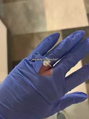 A hand wearing a blue glove is holding up a finger with a large diamond ring on it. The background shows a bathroom-like tile floor. A text overlay at the bottom says, "my wife is a doctor".