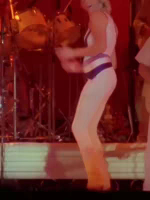 The image shows a person in a white outfit, likely a bodysuit or leotard, with a dark purple band around the waist. The person is in motion, mid-dance or performance, with their left arm up near a drum kit in the background. The lighting is warm with shades of red and orange.