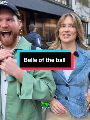 Two people, a man and a woman, are standing side-by-side outdoors. The man, on the left, is wearing a black baseball cap and a green jacket, with his mouth open in a surprised or excited expression. The woman, on the right, is wearing a blue denim jacket and has a neutral facial expression. A text overlay in a rectangular box reads "Belle of the ball", with the word "CUTE" in the lower-left corner, suggesting a romantic context.