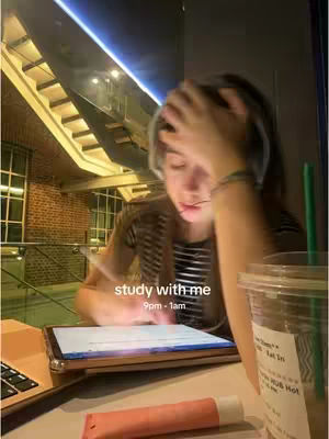A person wearing a striped shirt is sitting at a table, resting their hand on their head while they look down at a tablet. A laptop and a Starbucks cup are on the table with a tube of lipgloss. The background includes a window, brick wall, and stairs.